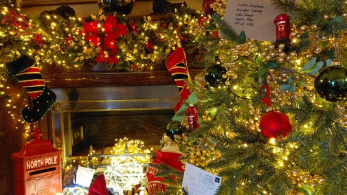 Decorated Christmas tree beside a fireplace, illuminated with warm white lights and adorned with red and black baubles, gold berry garlands, stockings, wrapped presents, and letters to Father Christmas, creating a festive holiday display.